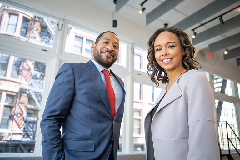 An African-American male and female smiling and wearing business suits. They are standing in the lobby of a business.
