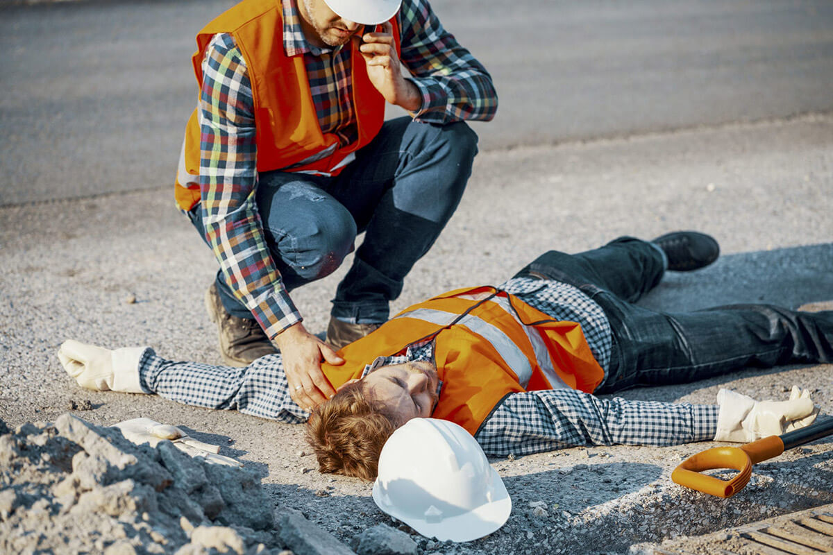 A construction worker lays on the ground, unconsious. A second worker kneels over him on the phone.