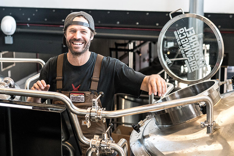 A smiling man wearing a hat and apron stands in front of open brewing equipment.