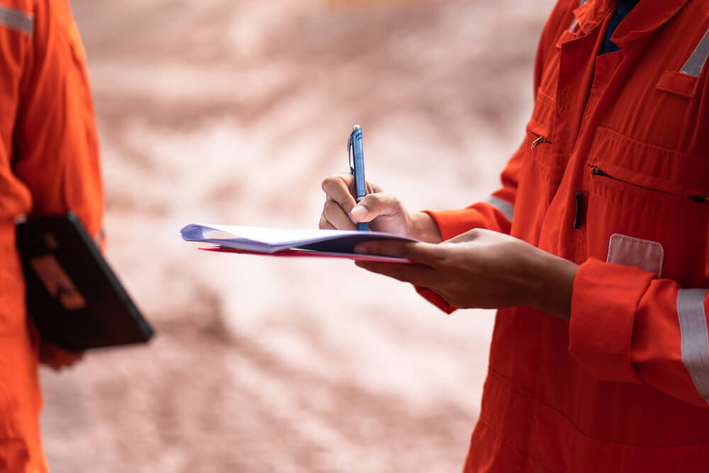 A worker in an orange jumpsuit stands with clipboard and pen in hand.